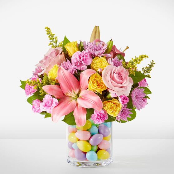 Spring flower arrangement with lilies, roses, carnations, daisy mums, and greenery in a glass vase filled with colorful Easter eggs