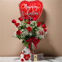 A dozen red roses with baby's breath in a vase, with a heart shaped balloon, stuffed animal and box of chocolates on a table.