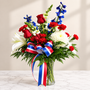 A patriotic red, white, and blue floral arrangement featuring red roses and carnations, white lilies and chrysanthemums, blue delphinium-style flowers, and lush green foliage. The bouquet is arranged in a clear glass vase and accented with a red, white, and blue striped ribbon, creating a bright, elegant design ideal for Memorial Day, Independence Day, Veterans Day, military tributes, patriotic celebrations, and remembrance occasions.