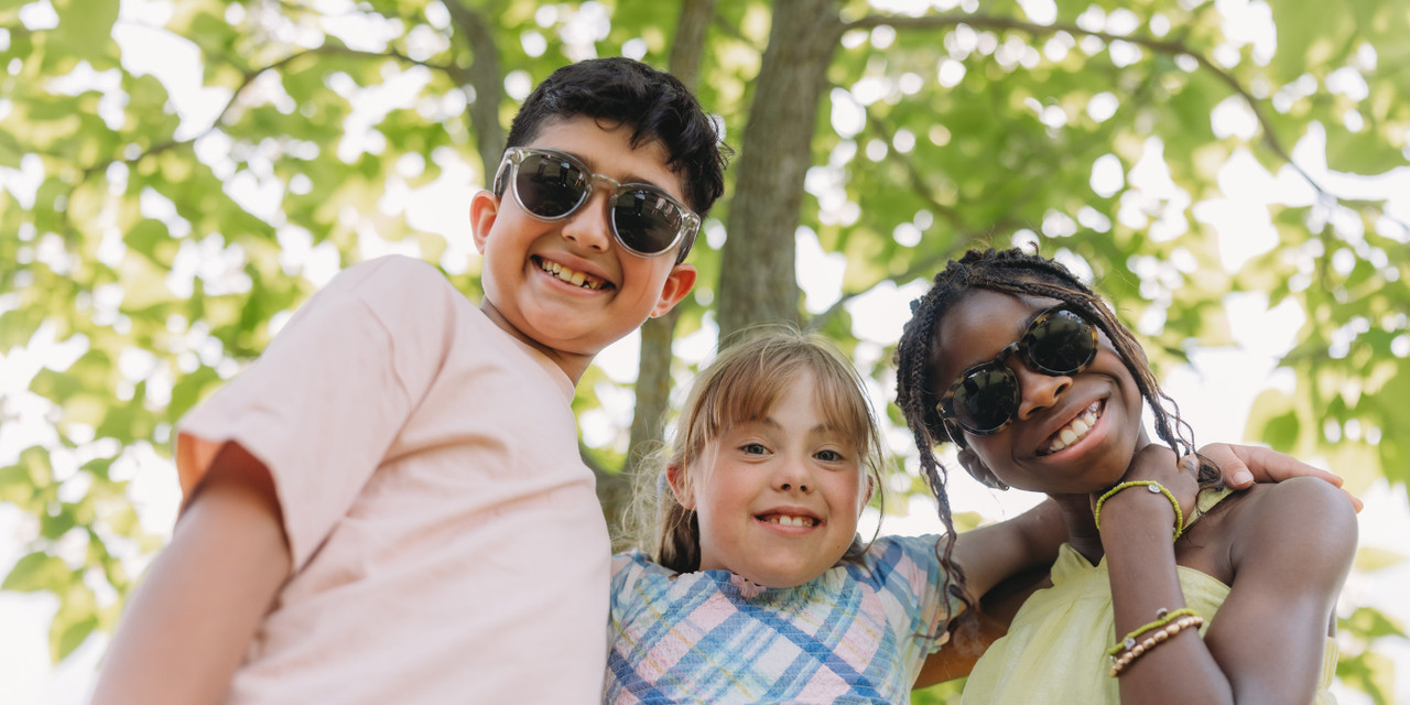 Three children wearing sunglasses stand close together outdoors, looking toward the camera with trees and green leaves overhead.