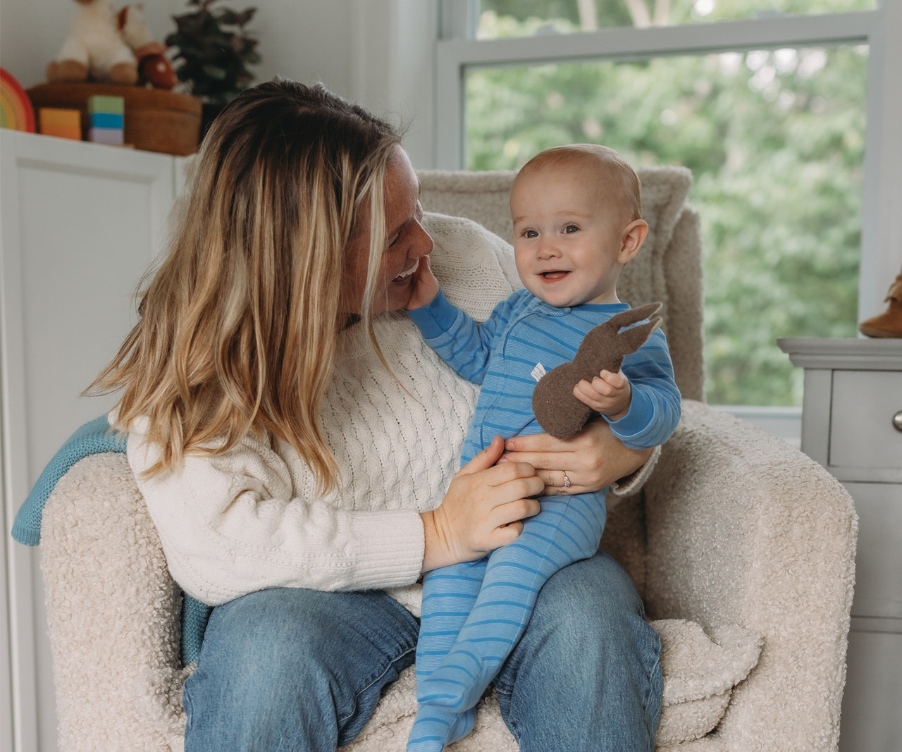 An adult sitting in a cozy chair holding a baby dressed in a blue striped onesie, who is holding a soft toy, in a bright living room.