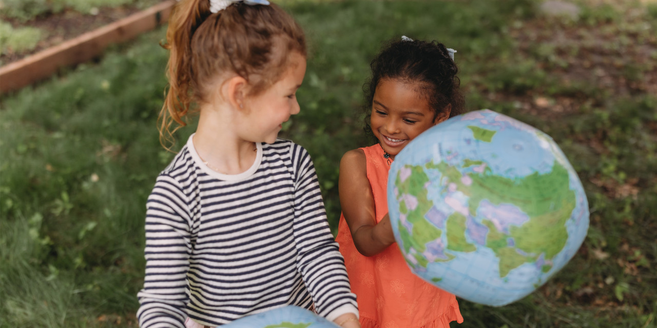 Two girls playing outdoors, each holding an inflatable globe ball, with grass and a wooden border visible in the background.