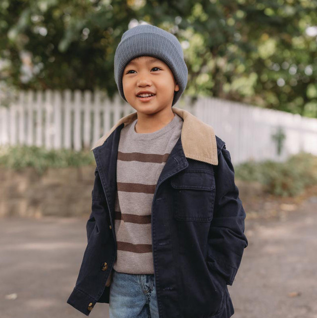 A boy wearing a gray beanie, striped shirt, and dark jacket with tan collar, standing outdoors near a white picket fence.
