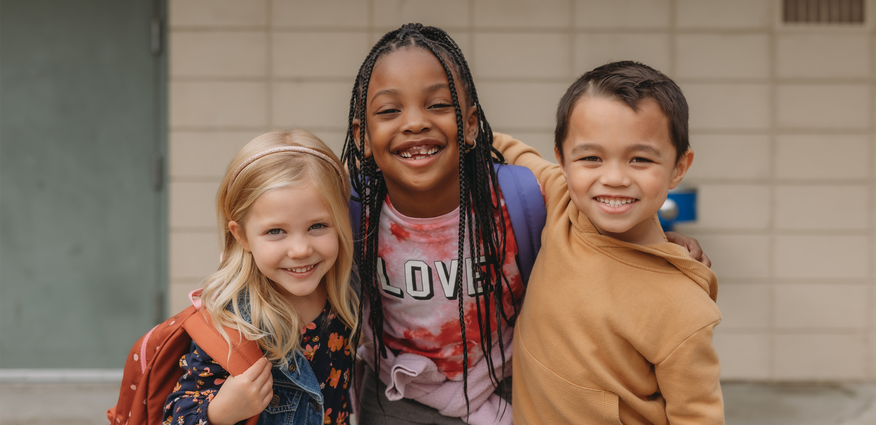 Three children standing close together outside a building with a beige brick wall, wearing casual outfits and backpacks.