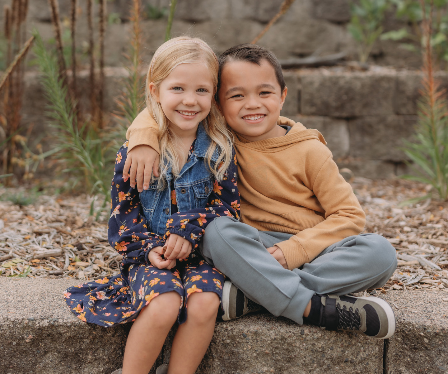 Two children sitting close together on a stone ledge outdoors, the girl is wearing a floral dress and the boy a tan hoodie.