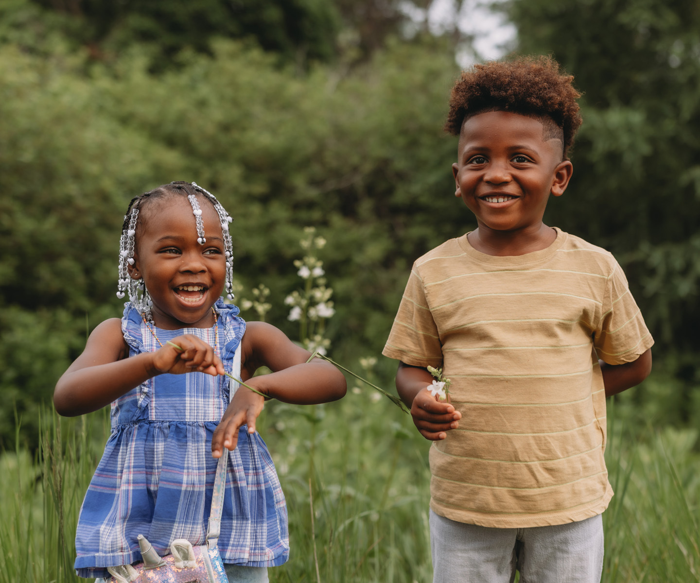Two children stand in tall grass outdoors, holding small wildflowers with trees in the background.