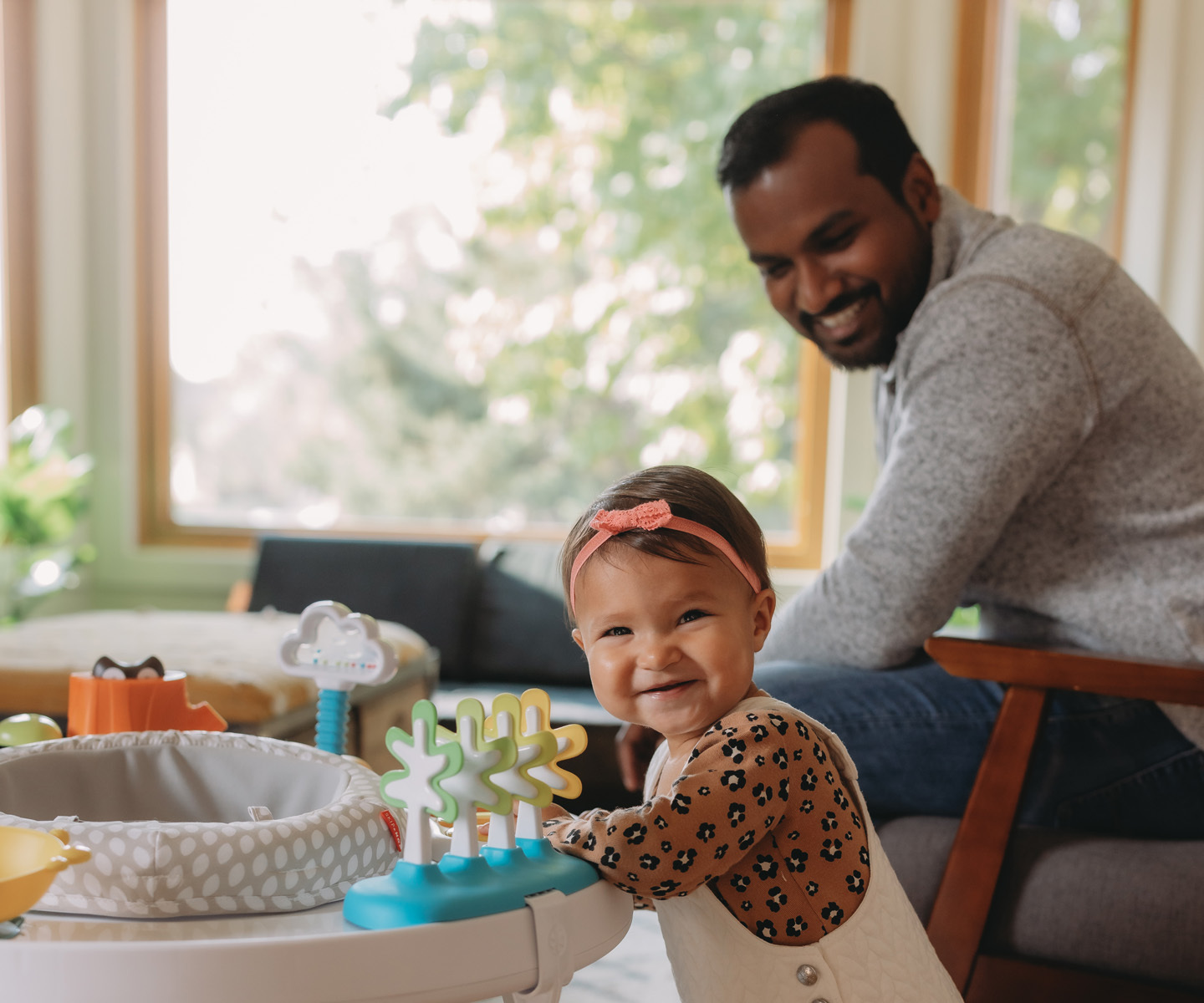A baby wearing a patterned outfit and pink headband stands at a colorful activity center, with an adult seated nearby in a bright living room.