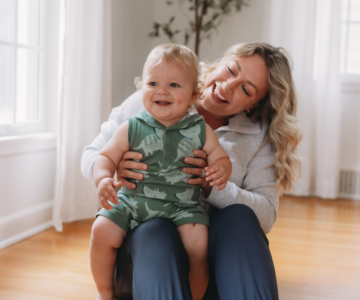 An adult sits on a hardwood floor holding a baby indoors, with soft natural light coming through nearby windows.