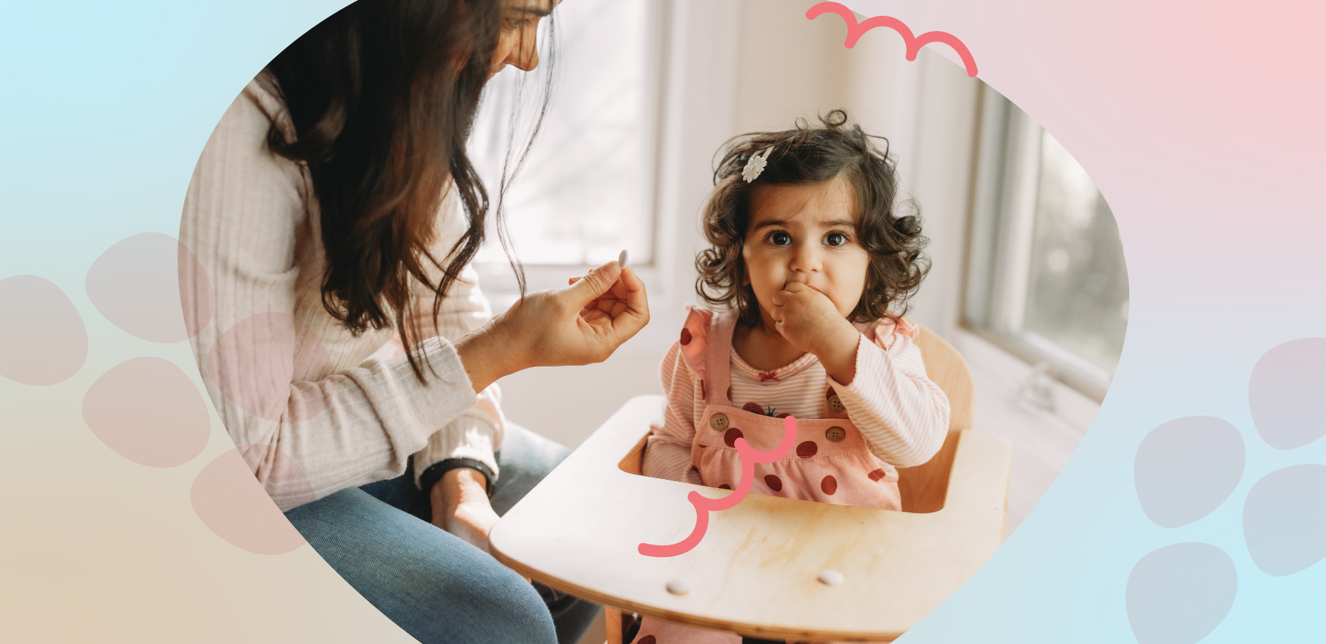 The image shows a woman with brown hair holding a little girl with brown hair and a dress. They are standing in a field of tall grass and flowers. The woman is smiling, and they are both looking down at the pink and white flowers in front of them, towards the bottom right of the image.