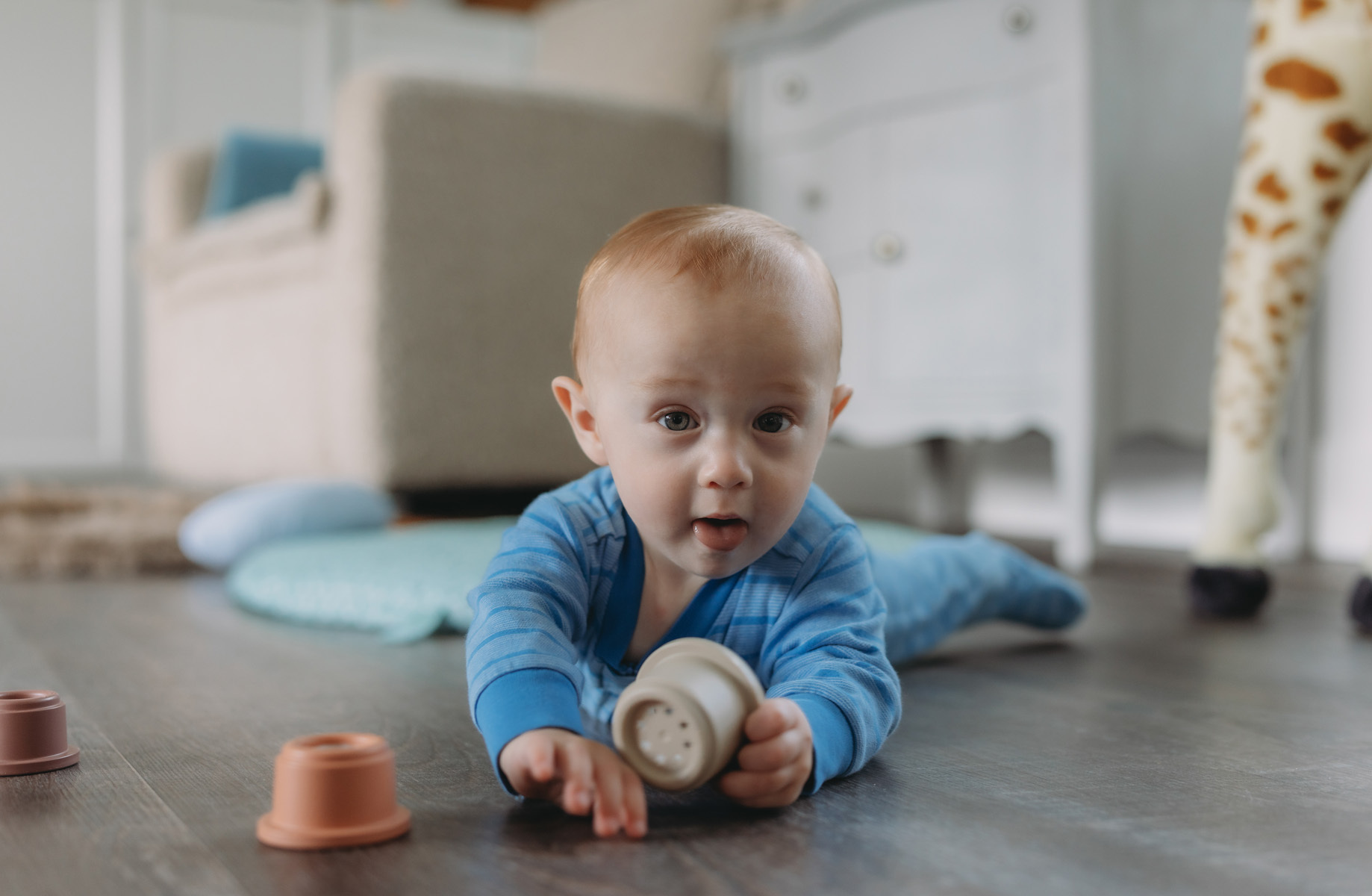 A baby in a blue onesie lying on a wooden floor, holding a small toy, with other toys scattered nearby in a cozy living room.