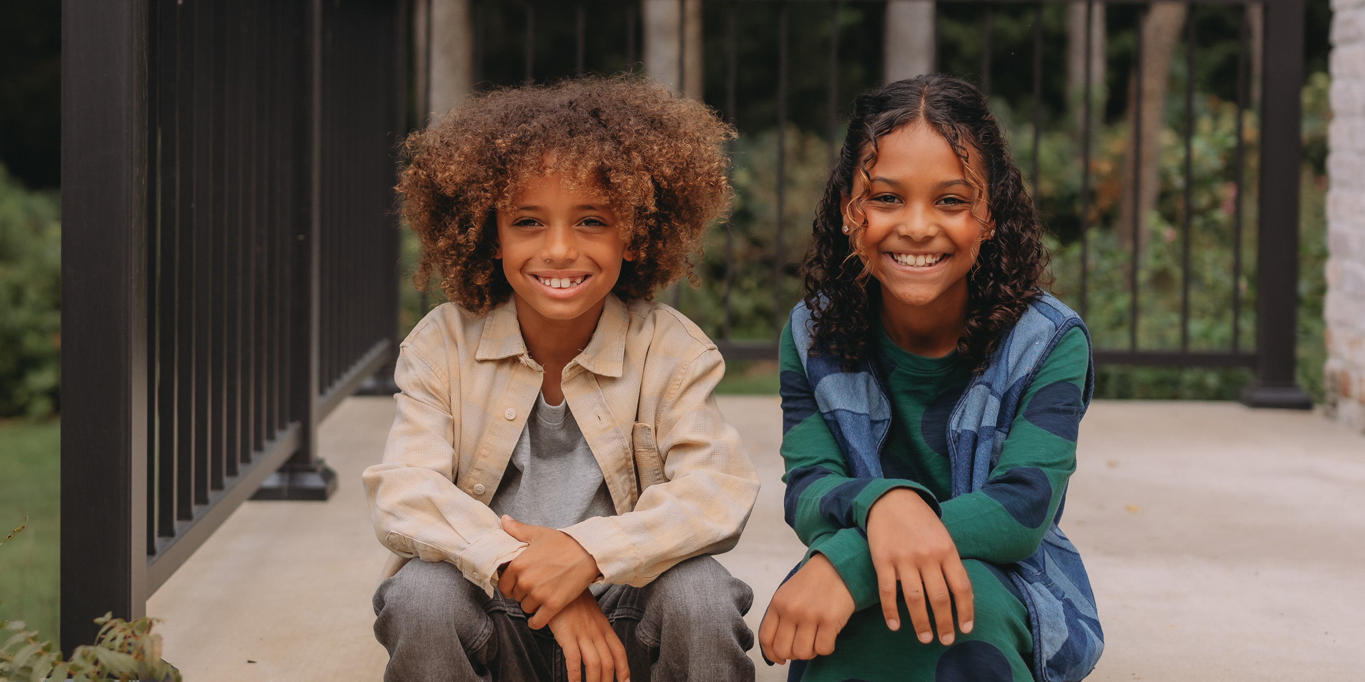 A boy and a girl sitting side by side on a porch with a black railing, wearing layered outfits in neutral and green tones.