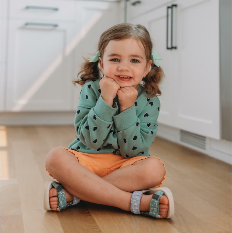 A young girl sitting on the kitchen floor, looking up with a smile