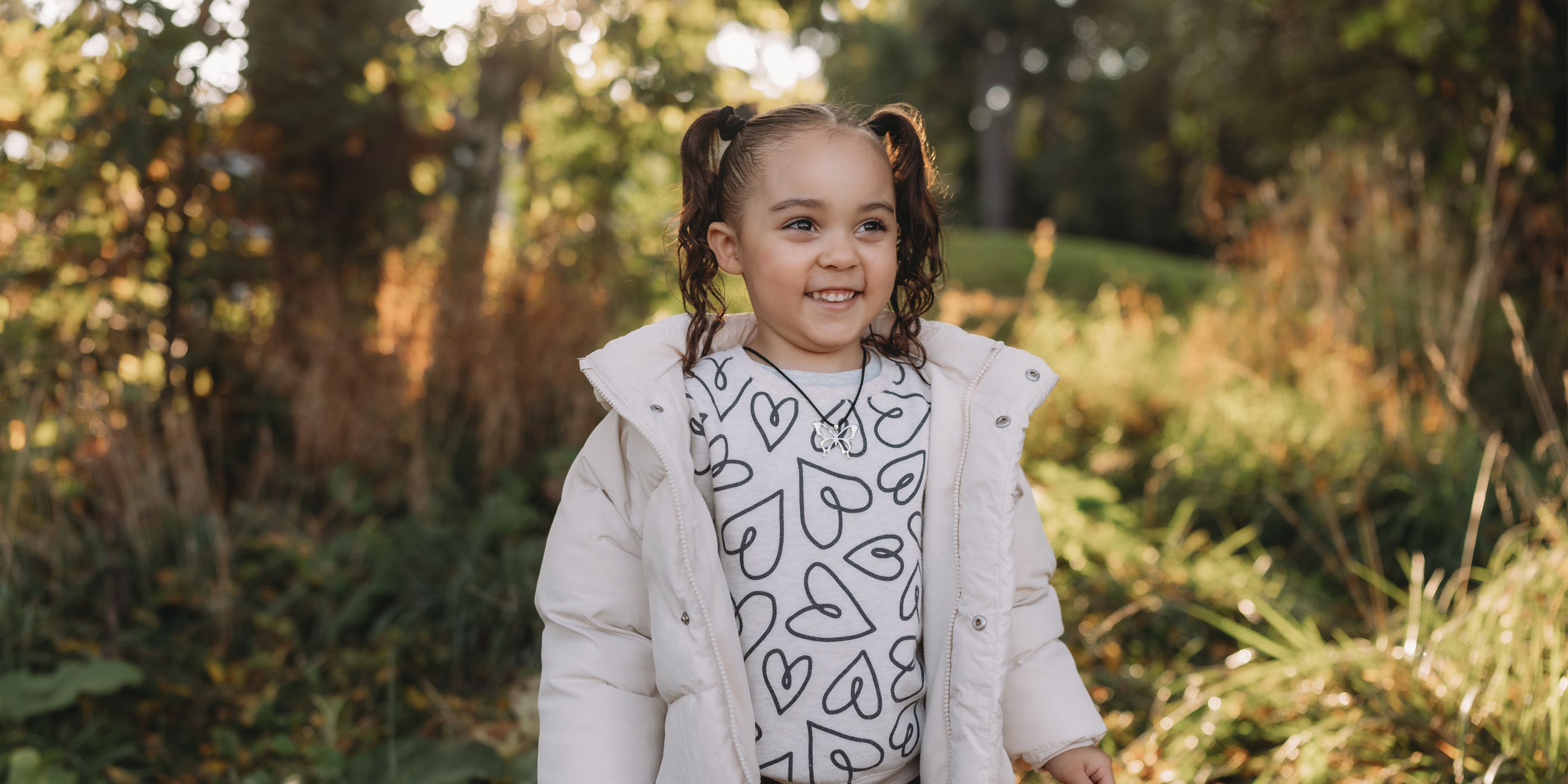 A girl wearing a white puffer jacket and a shirt with a heart pattern, standing outdoors in a grassy area with tall plants in the background.