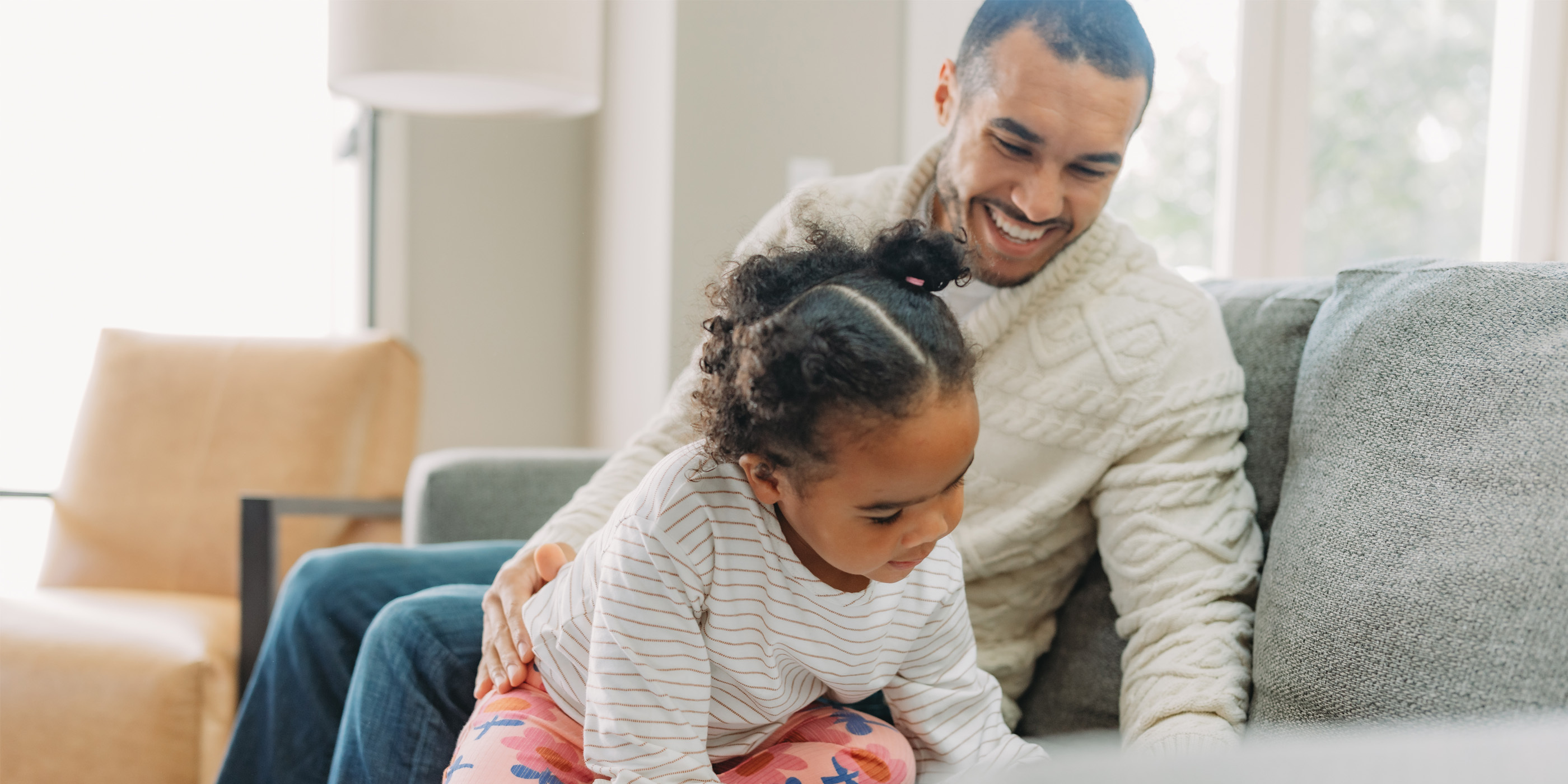An adult sitting on a couch with a child in a striped shirt and colorful pants leaning forward, in a bright living room setting.