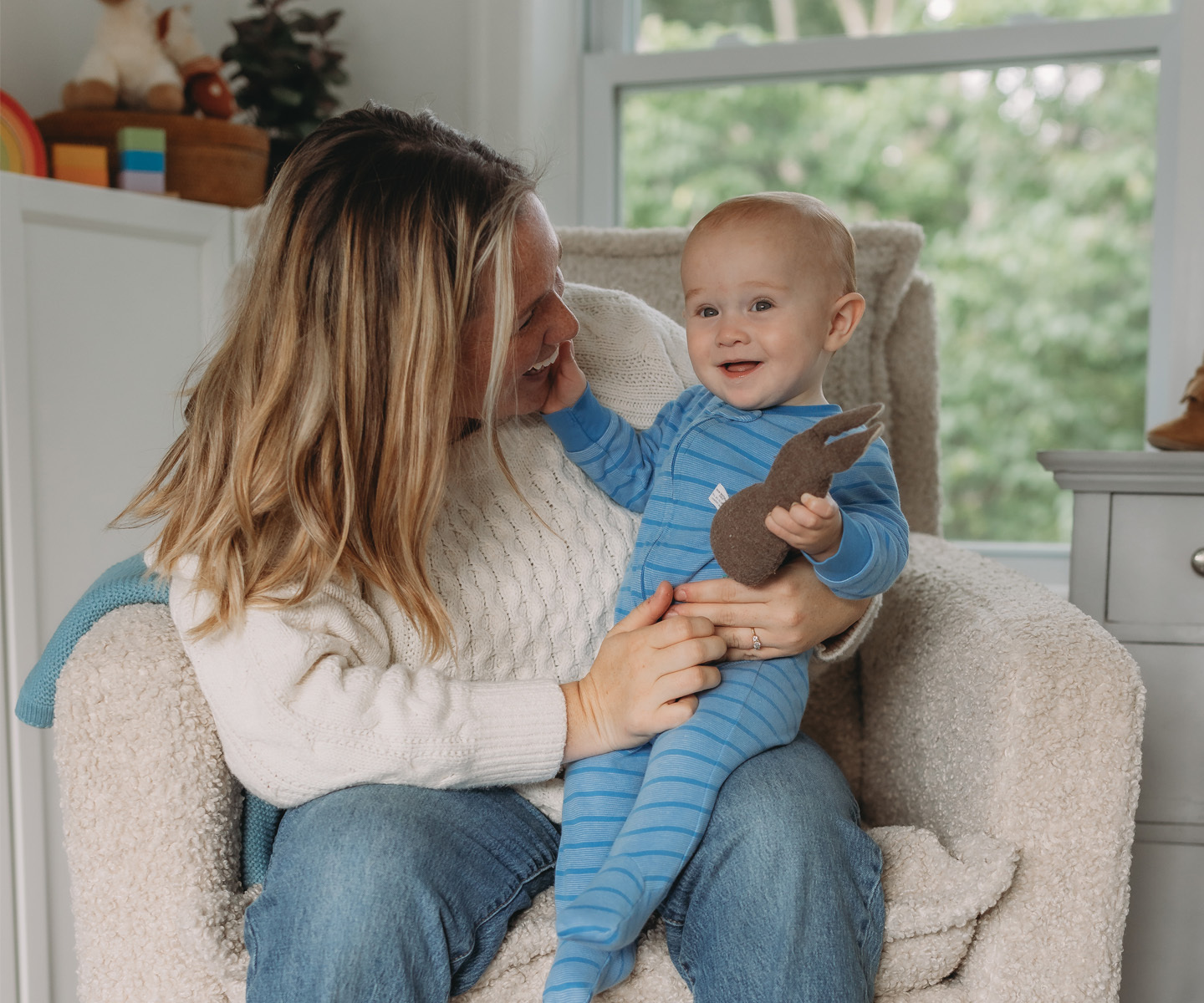 An adult sitting in a cozy chair holding a baby dressed in a blue striped onesie, who is holding a soft toy, in a bright living room.
