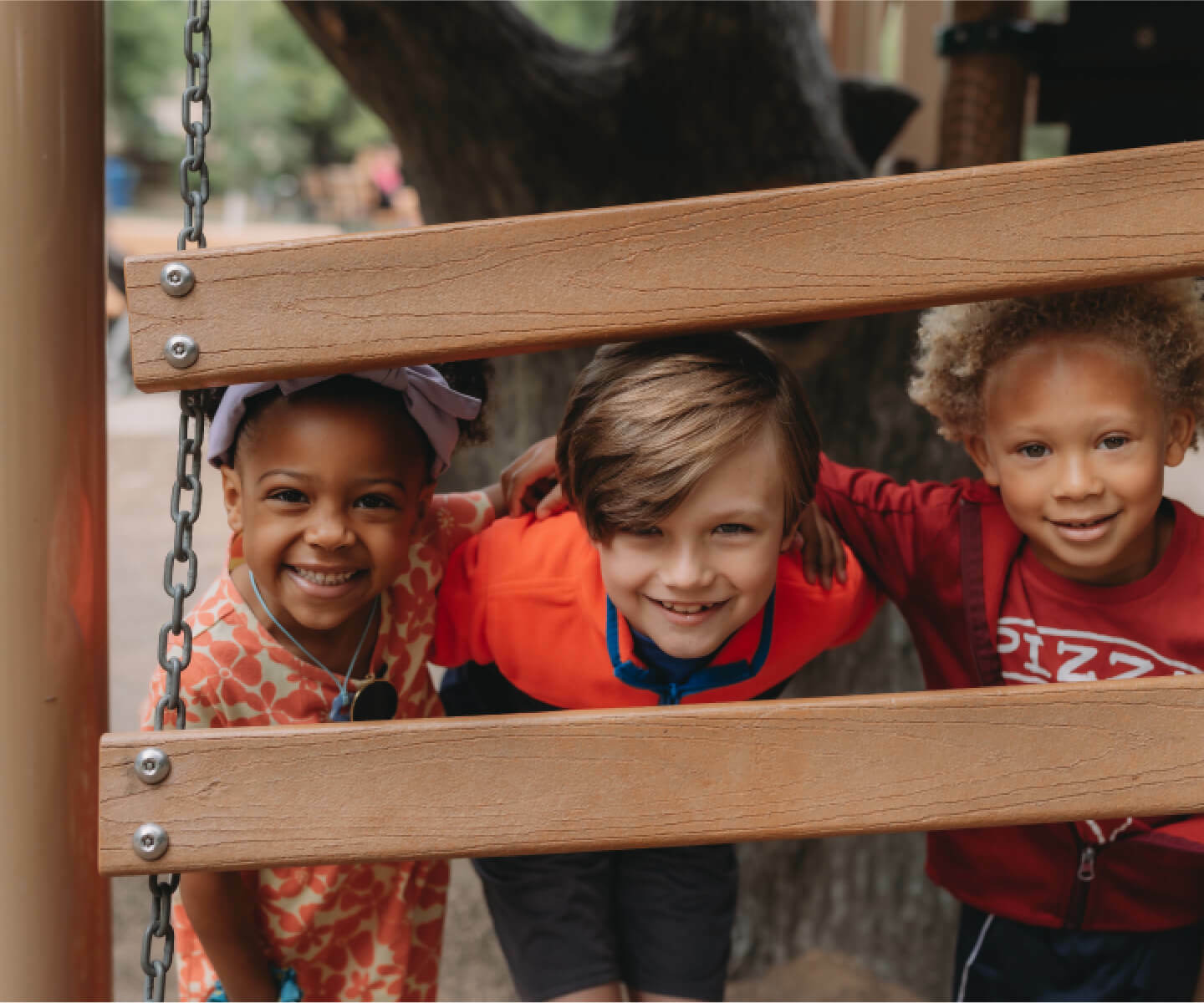 Three happy children standing on a playground and peeking from between two wooden ladder.