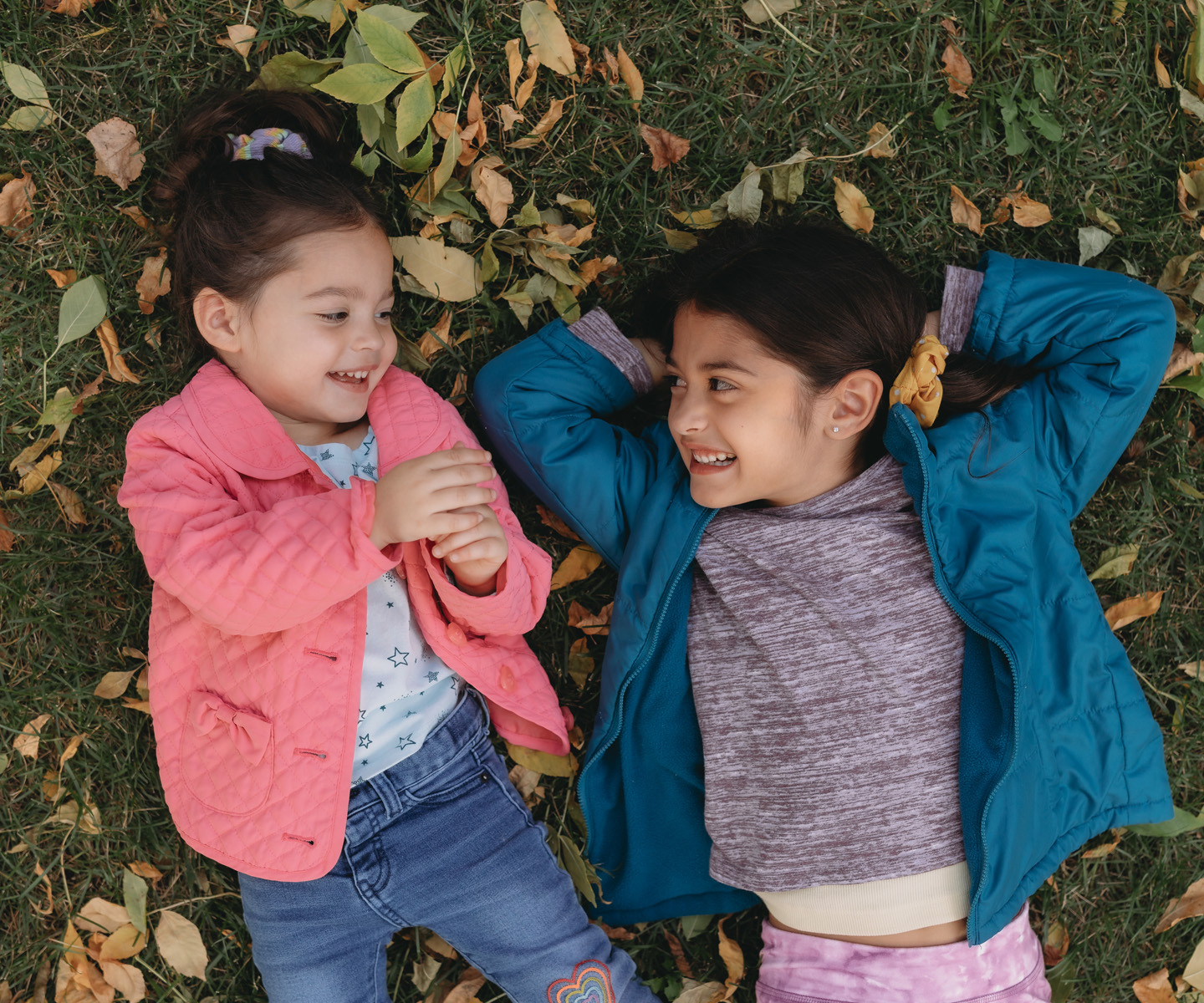 Two girls lying on grass covered with fallen leaves, wearing colorful jackets—one pink and one teal—engaged in a relaxed moment outdoors.