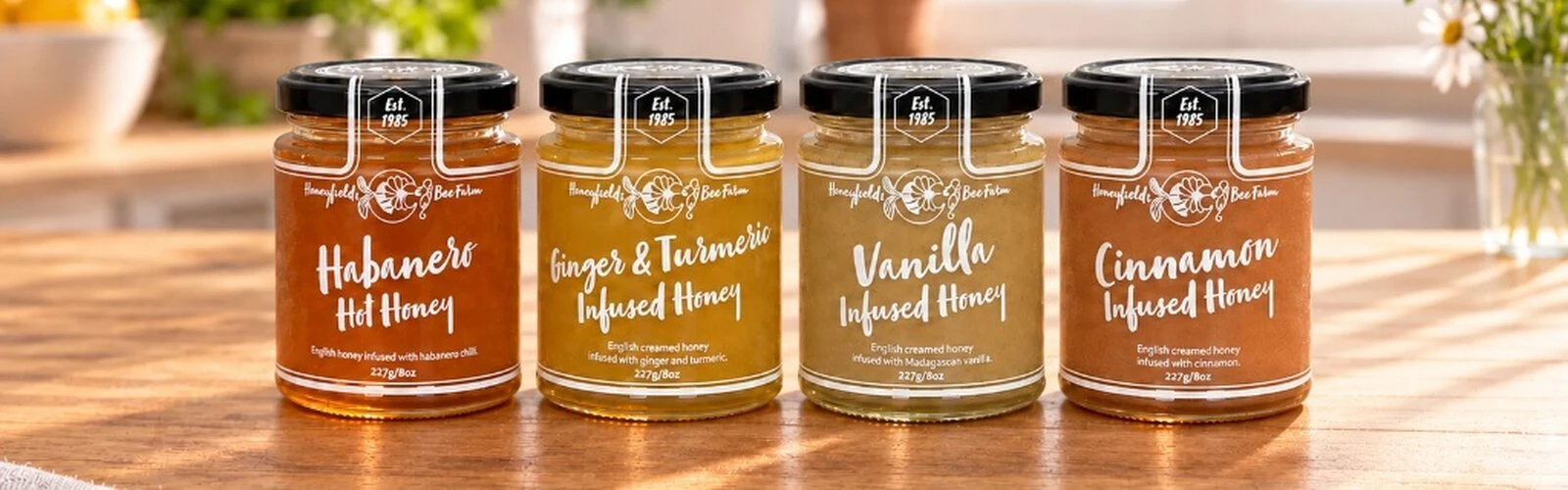 Four glass jars of Honeyfields Bee Farm Infused English Honey: Habanero, Ginger, Vanilla, and Cinnamon, on a rustic sunlit wooden table