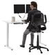 Sit-and-stand typist chair upholstered in black fabric without arms, at a computer desk.