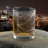 Elegantly engraved whiskey glass featuring a detailed map of Pittsburgh, resting on a stone surface with the sparkling cityscape and the illuminated bridges over the Allegheny River at night in the background, a thoughtful gift for University of Pittsburgh students or alumni.