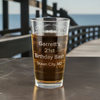 Custom laser etched pint glass featuring optional backside text. Ideal for personalized birthday celebrations, displayed with a scenic ocean background. Great for milestone birthday gifts and party favors.