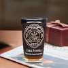 Custom engraved firefighter pint glass with Maltese Cross, featuring name and text. Displayed on a table with a wrapped gift and newspaper in the background, perfect as a thoughtful gift for firefighters and special occasions.