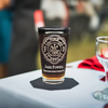Personalized firefighter pint glass engraved with Maltese Cross, name and fire company. Displayed on an event table with blurred guests in the background. Perfect for celebrations and gifts to honor firefighters at special events.