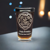 Personalized firefighter pint glass with laser-etched Maltese Cross and two lines of text. Displayed against a dark backdrop with soft blue and orange bokeh lights, highlighting its elegance and perfect for firefighter gifts or commemorative items.