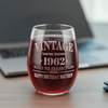 A personalized 15oz stemless wine glass filled with red wine, featuring the laser etched Vintage Aged to Perfection design with personalized year and message. Ideal for milestone birthday gifts. Glass is set next to a laptop.