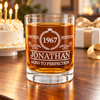 Personalized “Aged to Perfection” whiskey glass on a wooden table, with an ivory birthday cake, lit candles, and a wrapped gift in the background.