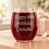 Backside of a stemless wine glass engraved “Happy 80th Birthday Grandma,” beside a blush-wrapped present.