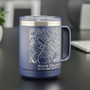 Blue stainless steel mug featuring a laser-etched map of State College, PA, showcasing precise street lines and local landmarks. Below the map, the coordinates 40°47'43.7\"N 77°51'41.8\"W and the text “STATE COLLEGE, PA” are engraved for added detail. The insulated 15oz travel mug is placed on a reflective surface, with a blurred background including a plant and a laptop, emphasizing its modern and functional design—ideal for home, office, or gifting.