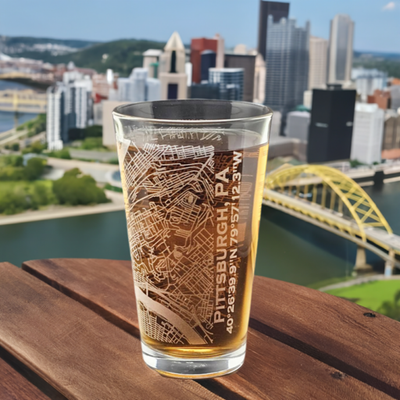 Laser etched pint glass with street level map of Pittsburgh, PA sitting on table overlooking the city skyline