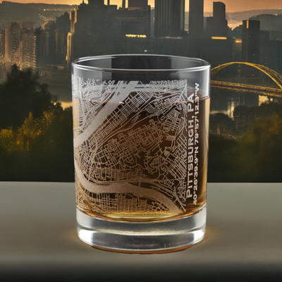 Elegant whiskey glass with intricate Pittsburgh city map engraving, positioned against an early morning cityscape with the iconic yellow bridge, ideal for collectors and as a sophisticated Pittsburgh souvenir.