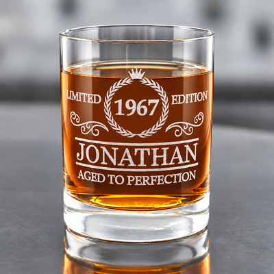 Clear heavy-base whiskey glass filled with amber liquid, featuring a personalized name and year within a crown-and-wreath “Aged to Perfection” design, on a gray surface.