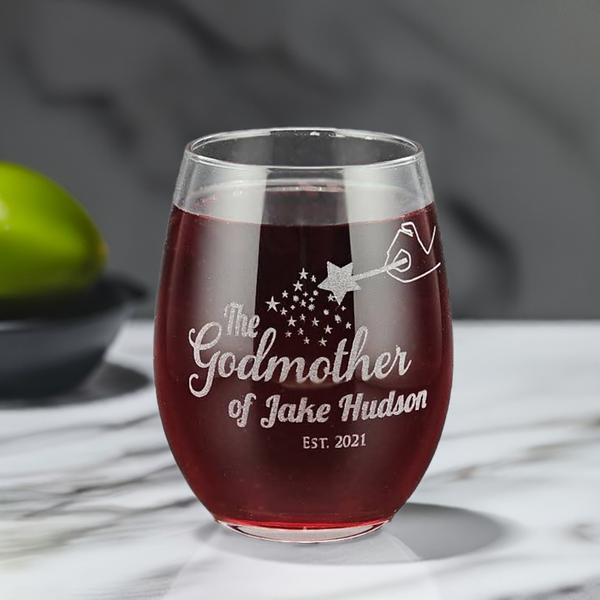 Personalized 15oz stemless wine glass with laser etched fairy godmother design, personalized with godchild name and date. Filled with red wine and placed on a marble surface, this custom glass makes a perfect gift for godmothers on special occasions.