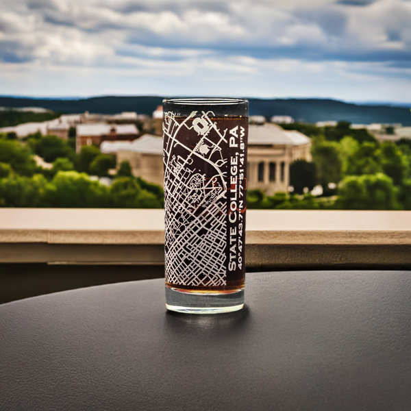 Highball glass with etched street map of State College, PA, displayed on an outdoor ledge with a view of Penn State University in the distance, perfect for graduation and alumni gifts