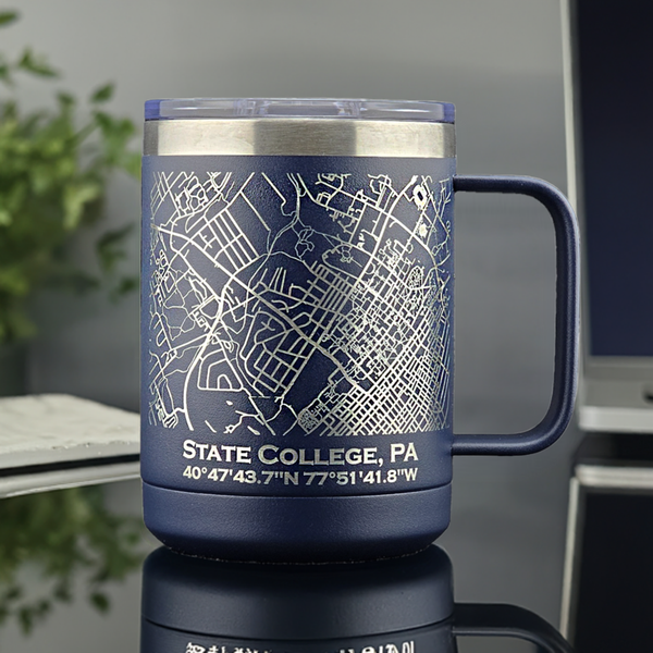 Blue stainless steel mug featuring a laser-etched map of State College, PA, showcasing precise street lines and local landmarks. Below the map, the coordinates 40°47'43.7\"N 77°51'41.8\"W and the text “STATE COLLEGE, PA” are engraved for added detail. The insulated 15oz travel mug is placed on a reflective surface, with a blurred background including a plant and a laptop, emphasizing its modern and functional design—ideal for home, office, or gifting.