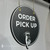 Side-angle view of a custom round ORDER PICK UP hanging sign featuring white vinyl lettering, a black PVC board, gold grommets, and decorative pointing hand.