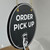 Angled product view of a black circular ORDER PICK UP business sign, highlighting its thick PVC board construction, white lettering, and gold hanging hardware.