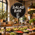 Custom round SALAD BAR hanging sign with white lettering and decorative pointing hand displayed above a fresh salad counter in a warm, rustic restaurant interior.