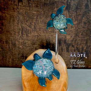 Flameworked glass art sculpture by AAOTE's founder, Tari Zarka. Totally one-of-a-kind, investment-quality glass art on a wooden base. 

This Sea Turtle mama and baby combo in turquoise whirling skies design is gorgeous. Mama includes dichroic glass in the shell body. It's a 3-piece set including the wood base. The overall size is approximately 7.75"H x 4"W x 5"D.

There is only one available. The piece you see is the piece you get. It will arrive dissassebled. The glass pieces sit on the base, and gets wrapped separate from the base for safe shipment. Assembly is as easy as placing the glass onto the wooden base. 

The wooden base is actually crafted from fallen timber from AAOTE's property. 

This sculpture will ship USPS Priority with insurance and is not eligible for free shipping offers.