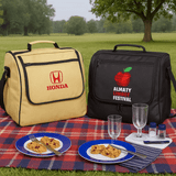 Two picnic bags, one yellow and one black, on a picnic blanket with plates of food and glasses.
