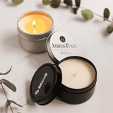 Three scented candles in silver and black containers, one with a lit wick, accompanied by green leaves.