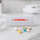 A transparent pill box with compartments for each day of the week, featuring various coloured pills beside it. It has a logo.