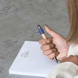 A wooden pen with a blue grip is being held over a blank notepad on a textured surface.