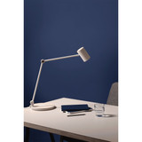 A white desk lamp with an adjustable arm, a blue notebook, and a glass of water on a light wood table against a blue wall.