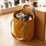 A yellow tote bag with grey straps, filled with towels, headphones, and a drink bottle, positioned near a table.