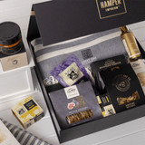 A gift pack featuring a grey blanket, gourmet snacks, nut mix, crackers, and a jar of preserve, all in a black box.