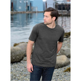 A man stands by a body of water wearing a dark grey short-sleeve t-shirt and jeans, with a rocky shoreline in the background.